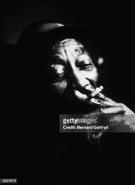 Headshot portrait of American blues musician Mississippi John Hurt smoking a cigarette.