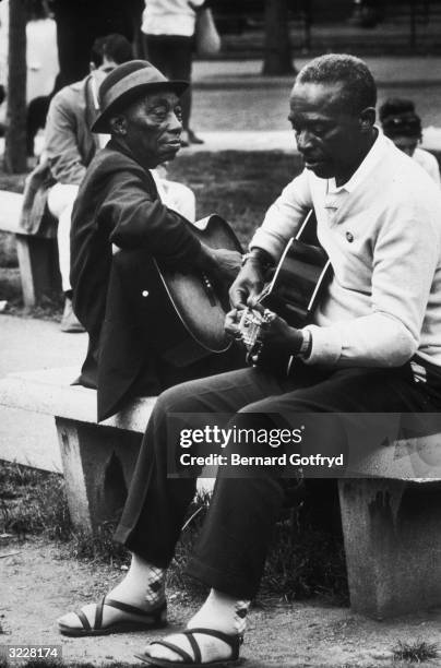 American blues musicians Mississippi John Hurt and Skip James play guitar while sitting on a bench in Washington Square Park, New York City.