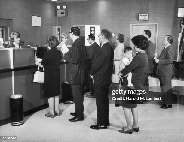Customers wait in line in front two tellers inside a bank, 1960s. A woman holds her baby at the end of the line.