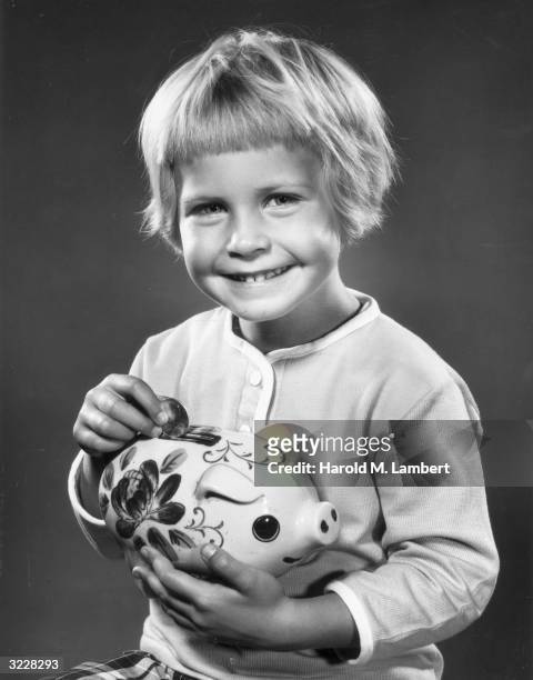Studio image of a young blonde girl smiling as she drops a silver dollar into her ceramic piggy bank, which she holds in front of her, 1960s.
