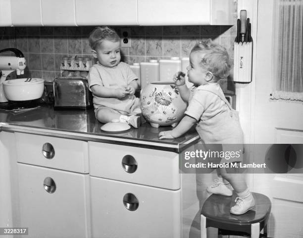 Pair of twins in matching overalls and T-shirts eat cookies taken from a ceramic container on a kitchen counter. One of them stands on a stool, while...