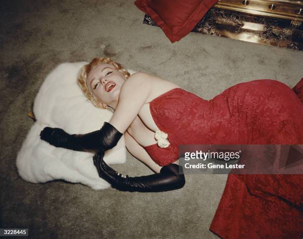 American actor Marilyn Monroe , wearing a red brocade evening gown and long black gloves, lying on a carpet and using a white fur stole as a pillow.