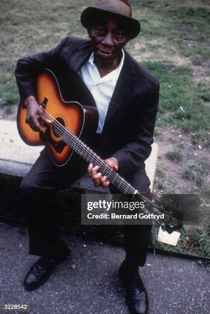 American blues musician Mississippi John Hurt plays a guitar while sitting on a bench in Washington Square Park, New York City.