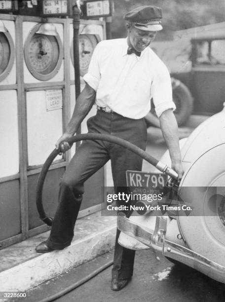 African-American athlete Jesse Owens fills up a car at a petrol station in his a uniform of cap, shirt and bow tie. Owens worked as a petrol pump...