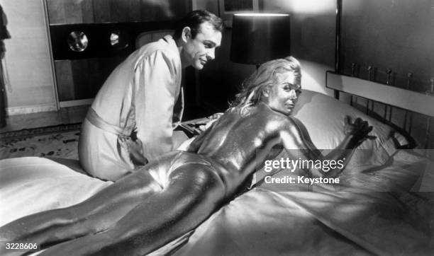 Sean Connery sitting beside his co-star English actress, Shirley Eaton, covered in gold during the filming of a scene from the James Bond film...