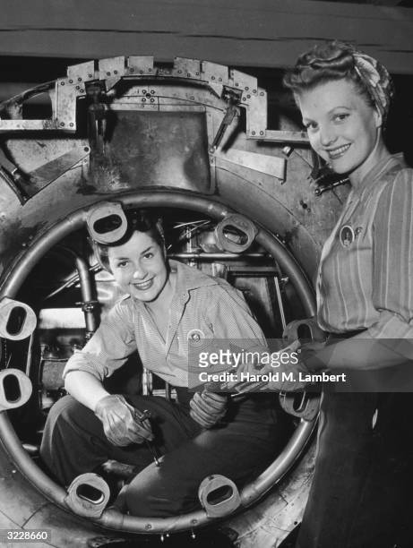 Two smiling women wearing union pins and checkered shirts hand each other tools during work in a factory during World War II.