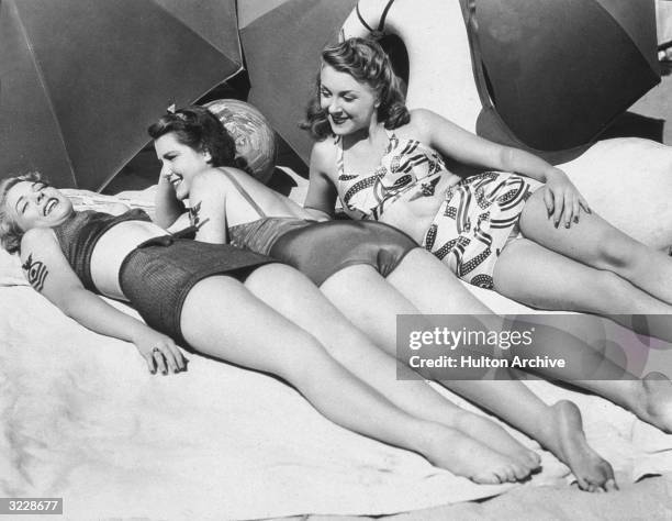 American actors Anne Gwynne, Evelyn Ankers, and Helen Parrish lay in the sun, attempting to create 'sun tattoos' of military insignia on their bodies...