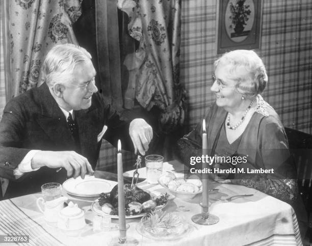 An elderly man and woman smile while sitting at a table having a candlelit dinner. The man is readying to carve the roast.