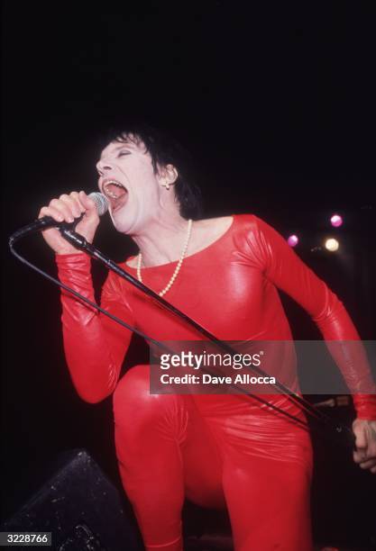 American rock singer Lux Interior of the group the Cramps crouches and sings into a microphone on stage during a performance. He wears a red vinyl...