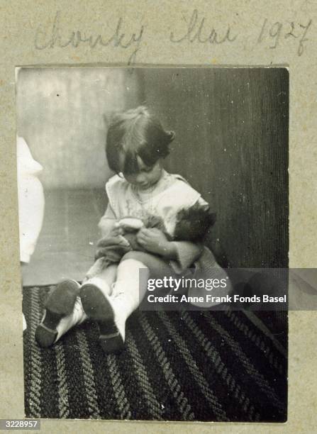 Photograph from Anne Frank's photo album of an unidentified young girl sitting on a rug and playing with a doll on her lap, with 'Chonky' and the...