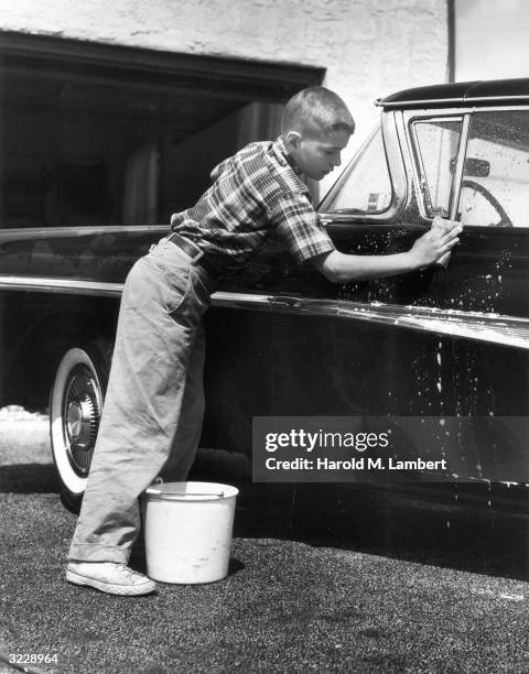 Boy with a crewcut washes the side of a Ford Fairlane automobile with a soapy sponge and a bucket of water in the driveway of his home, 1960s.