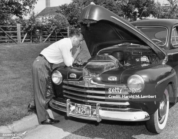 Man looks under the hood of his Ford and tries to figure out how to fix the engine after a breakdown on the side of the road, 1940s. He wears...