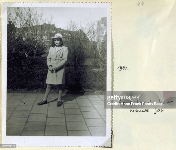 Full-length portrait of Anne Frank wearing a hat and coat standing on a sidewalk in front of shrubbery taken from her photo album, Amsterdam, Holland.