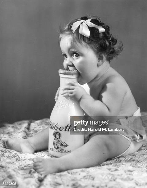 Studio image of an infant biting an Abbott's Ice Cream milk bottle, which sits between her legs. She wears a ribbon in her hair and a cloth diaper.
