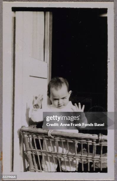 Anne Frank standing and waving her hands in the air in a wicker cradle on a balcony, Frankfurt am Main, Germany. From Anne Frank's photo album.