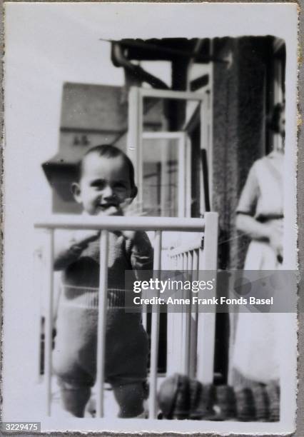 Anne Frank standing in a crib on a balcony as a woman looks on in the background, Frankfurt am Main, Germany. From Anne Frank's photo album.