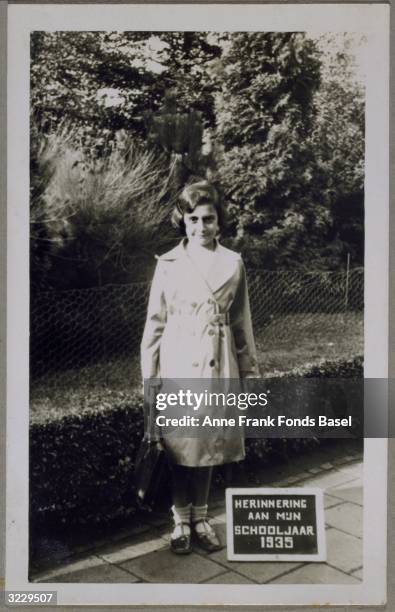 Full-length portrait of Margot Frank, the sister of Anne Frank, holding her school bag next to a plaque that says 'Memory of My School Year 1935' in...