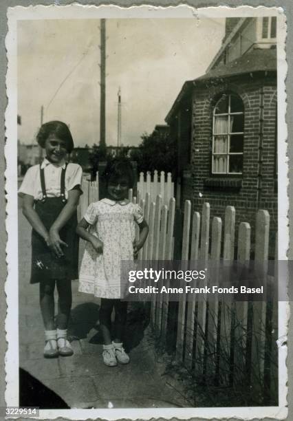 Full-length portrait of Anne Frank , and her sister Margot Frank standing on a sidewalk next to a fence. From Anne Frank's photo album.