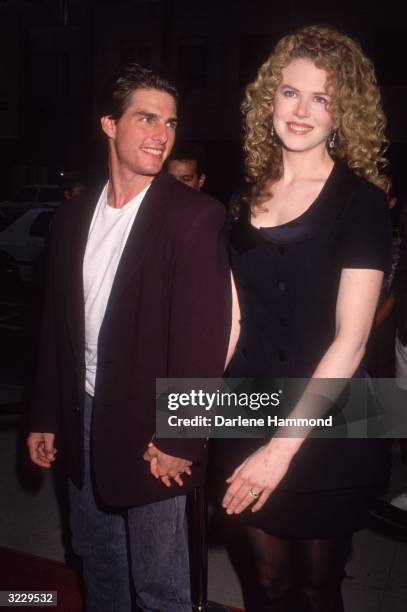 Married actors Tom Cruise and Nicole Kidman smile while holding hands on a red carpet. Cruise has short hair and wears a blazer with a white T-shirt...