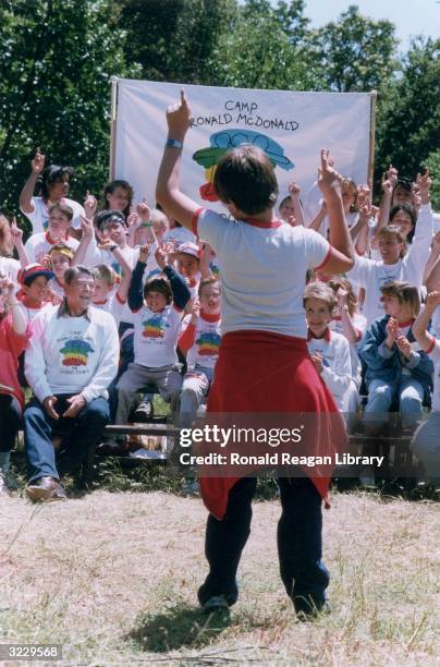 President Ronald Reagan and First Lady Nancy Reagan sit outdoors with a group of children, as a boy leads them in a song, at the Ronald McDonald Camp...