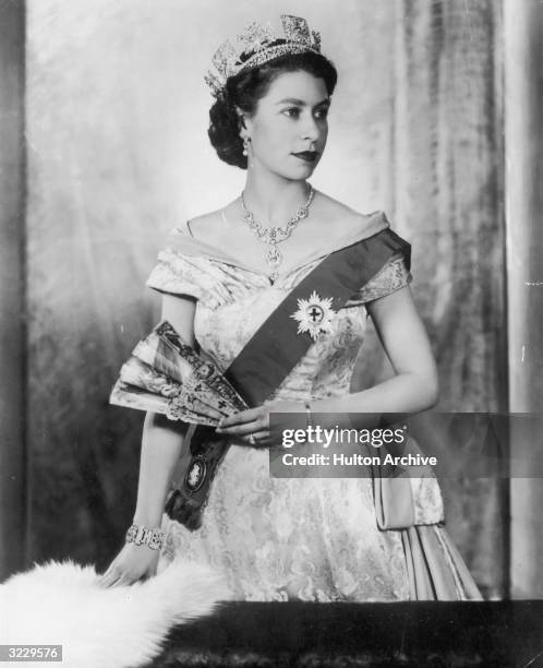 Studio portrait of Queen Elizabeth II holding a fan while wearing a brocade dress. She is also wearing a sash and the star of the Order of the...