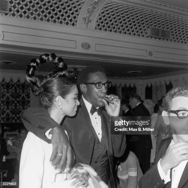 American comedian and actor Bill Cosby smokes a cigar while standing with his arm around his wife, Camille Hanks, at a Four Tops concert at the...