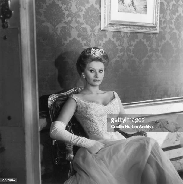 Italian actor Sophia Loren wears a ball gown, long white gloves, and a tiara while sitting in an armchair on the set of director Michael Curtiz's...