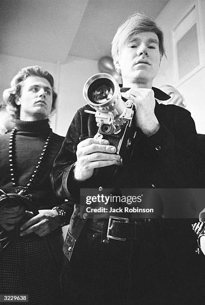 American pop artist Andy Warhol holds a camera fitted with a zoom lens while standing with photographer, poet, and archivist Gerard Malanga at club...