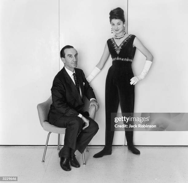 Italian fashion designer Emilio Pucci sits in a chair while a model wears one of his designs: a velvet jumpsuit with long white gloves.