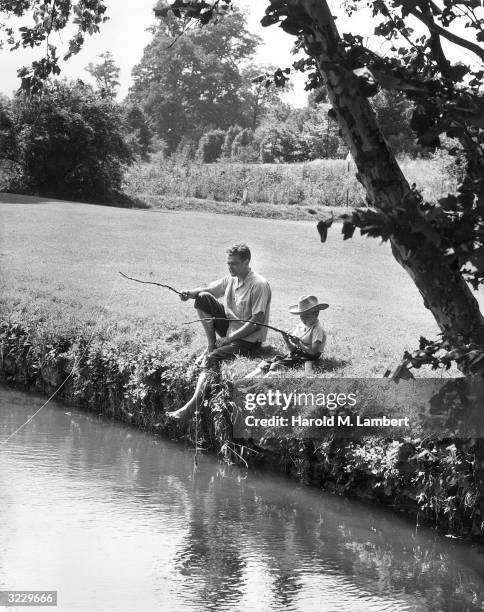 Father and his young son sit on the grassy banks of a river, fishing with homemade stick fishing poles, 1950s. Both are barefoot and the boy wears a...