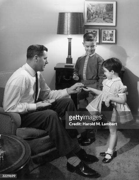 Father sits in an armchair in his living room, handing change to his son and daughter, 1950s. The girl smiles and holds out her hand.
