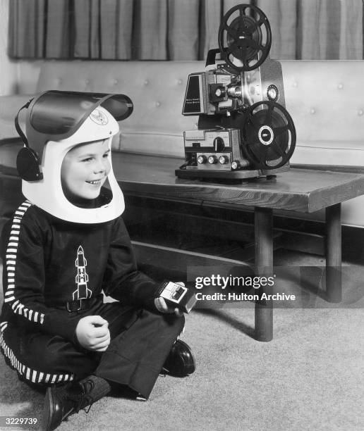 Boy wearing a space helmet and costume watching a film on his Bell & Howell Wireless Tele-Sonic 8mm motion picture projector, 1950s. He is holding...
