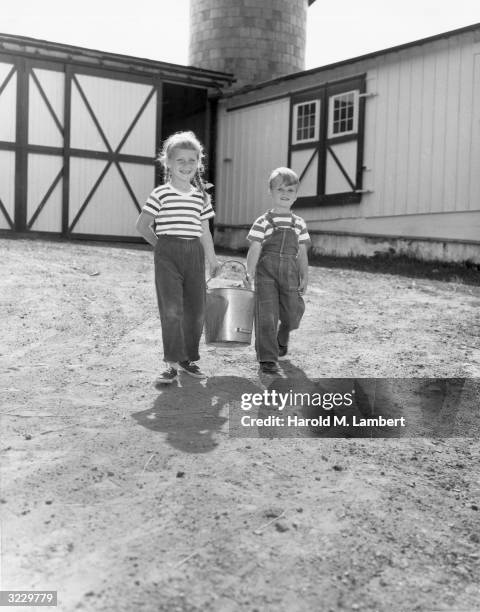 Young girl and boy smile as they walk in front of a barn, carrying a metal milk pail between them. Both wear striped T-shirts.