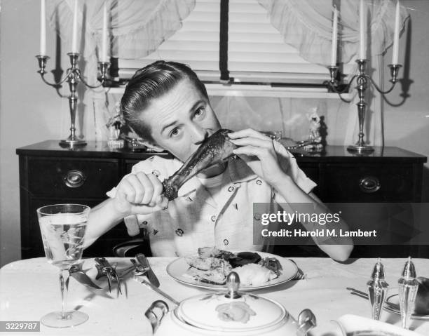 Teenage boy tilts his head to eat a drumstick while sitting with his elbows propped on the dinner table. The rest of his Thanksgiving meal consists...