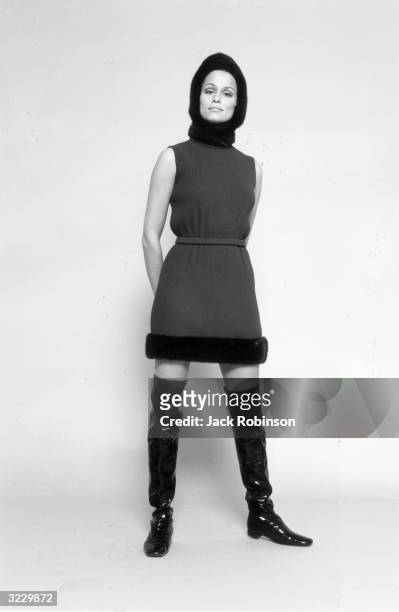 Full-length studio portrait of American model and actor Lauren Hutton standing in a wool mini-dress with fur trim, a fur hat, and thigh-high patent...