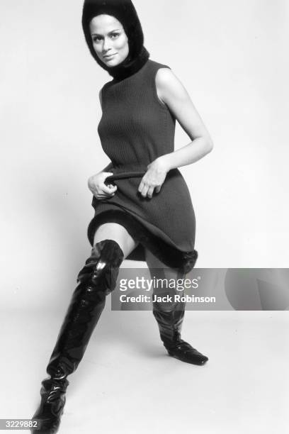 Full-length studio portrait of American model and actor Lauren Hutton, wearing a wool mini-dress with fur trim, a fur hat, and thigh-high patent...