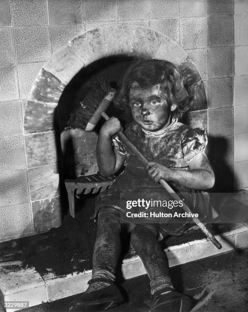 Young girl sits by a tile fireplace covered with soot, holding a chimney brush.