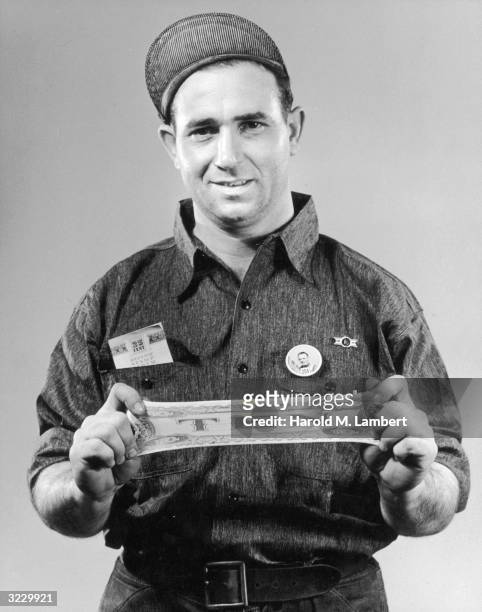 Studio portrait of a male worker standing and stretching a rubber ten dollar bill. He wears a cap and an inspection button on his shirt. He has a...