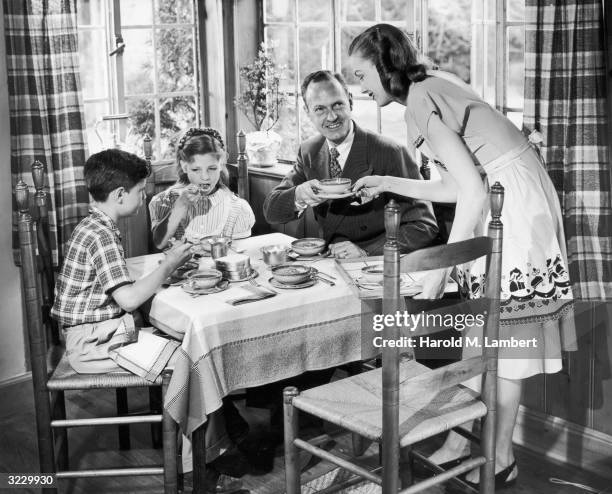 Housewife smiles as she hands a bowl of soup to her husband, who sits at the dinner table with their young son and daughter. She wears an apron.