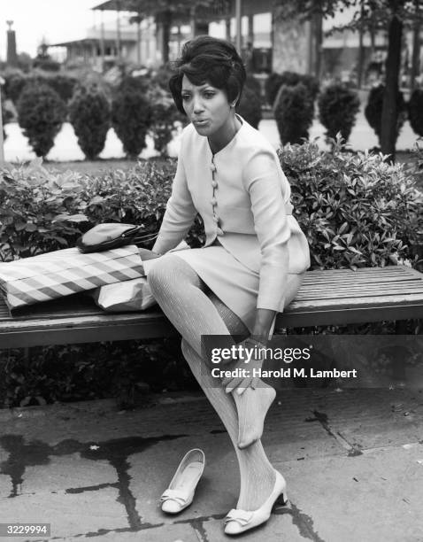 An African-American woman sits on a bench and rubs her ankle after removing her shoe. Her purse and shopping bags sit next to her. She wears a skirt...