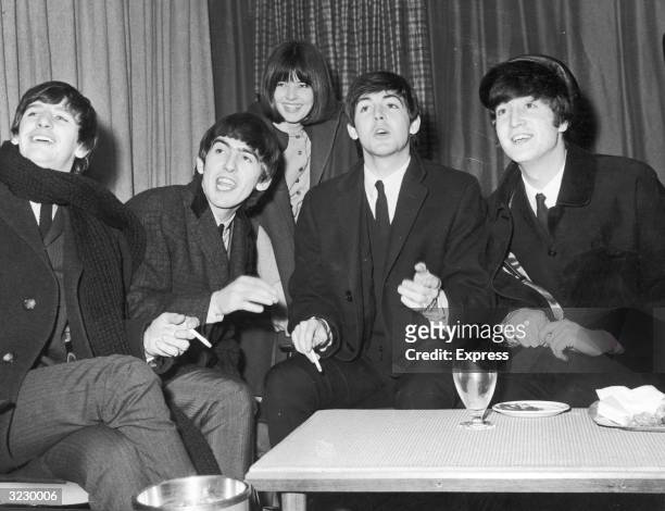 The British rock group The Beatles wear overcoats and smoke cigarettes while talking to reporters, including Maureen Cleave of the Evening Standard,...