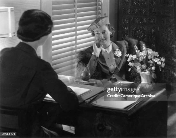 Woman sits at an office desk with her back to the camera, speaking to a woman on the opposite side of the desk. The woman wears a hat and a flower on...