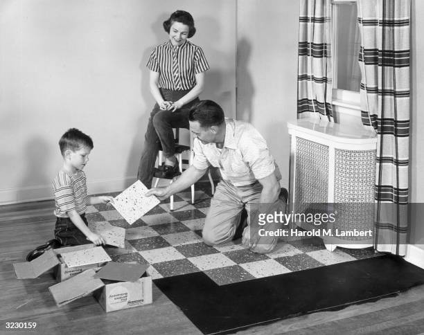 Father and son place linoleum tiles over a wooden floor in a living room while the mother watches from a stool.