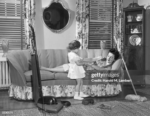 Housewife sleeps on the sofa while taking a break from her housework until her young tries to wake her. A vacuum cleaner and a dust broom sit by the...