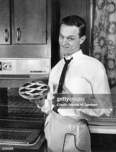 Young man smiles while holding a freshly baked cherry pie in front of an open oven in a kitchen. He wears an apron around his waist.