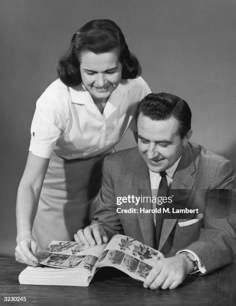 Studio image of a man sitting and looking through a women's fashion catalogue, as a woman stands behind him, smiling.