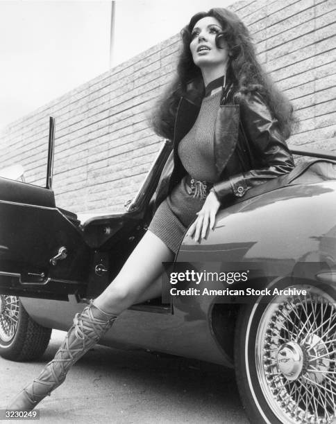 Low angle image of a woman stepping out of an E-Type Jaguar convertible sportscar, wearing a leather jacket, a minidress and laced cowhide boots.