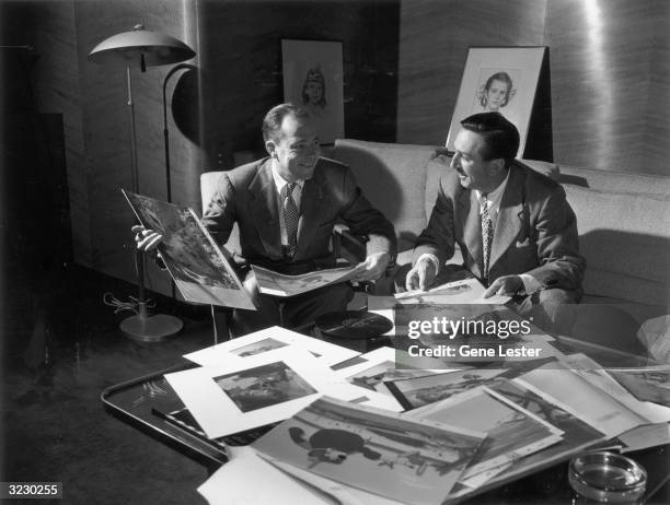 American animator and producer Walt Disney sits on a couch next to American songwriter Johnny Mercer in front of cartoon storyboard sketches posted...