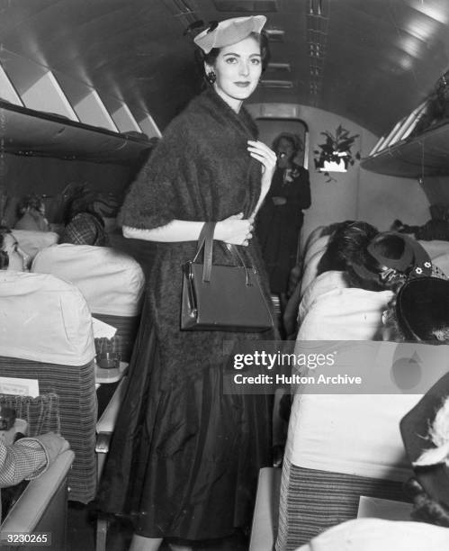 American model Carmen Dell'Orefice wears a hat and a mohair wrap and carries a purse while walking in the aisle of an airplane during a fashion show.