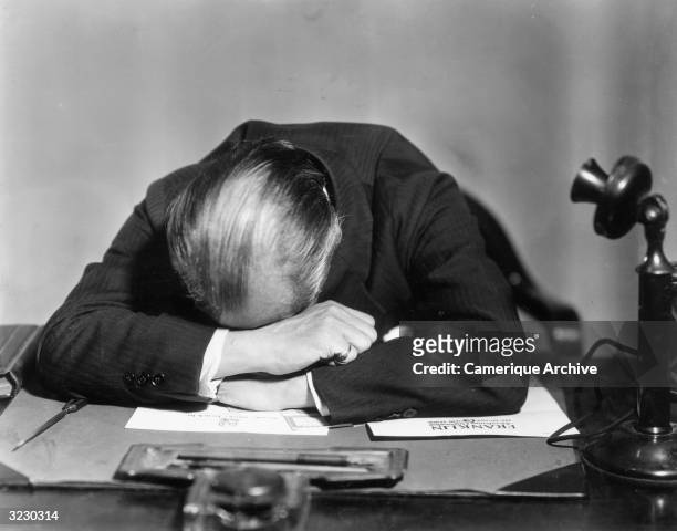 Businessman sits with his head face down on his desk. A telephone and some business documents sit on the desk.